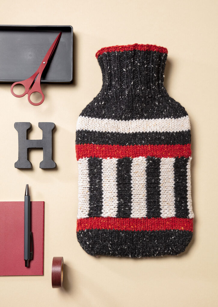 Produktfoto eines gestrickten Wärmflaschenbezugs mit Streifenmuster als Flatlay fotografiert mit schwarzen und roten Deko-Elementen