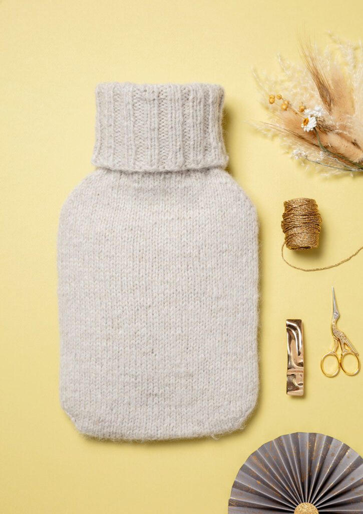 Produktfoto eines gestrickten Wärmflaschenbezugs als Flatlay fotografiert mit goldenen Deko-Elementen