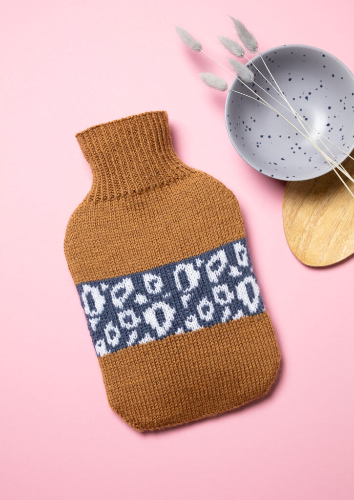 Produktfoto eines gestrickten Wärmflaschenbezugs mit Leo-Muster als Flatlay fotografiert mit braunen und blauen Deko-Elementen