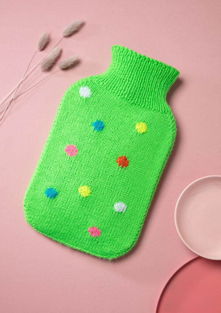 Produktfoto eines gestrickten Wärmflaschenbezugs mit bunten Punkten als Flatlay fotografiert mit rosa Deko-Elementen
