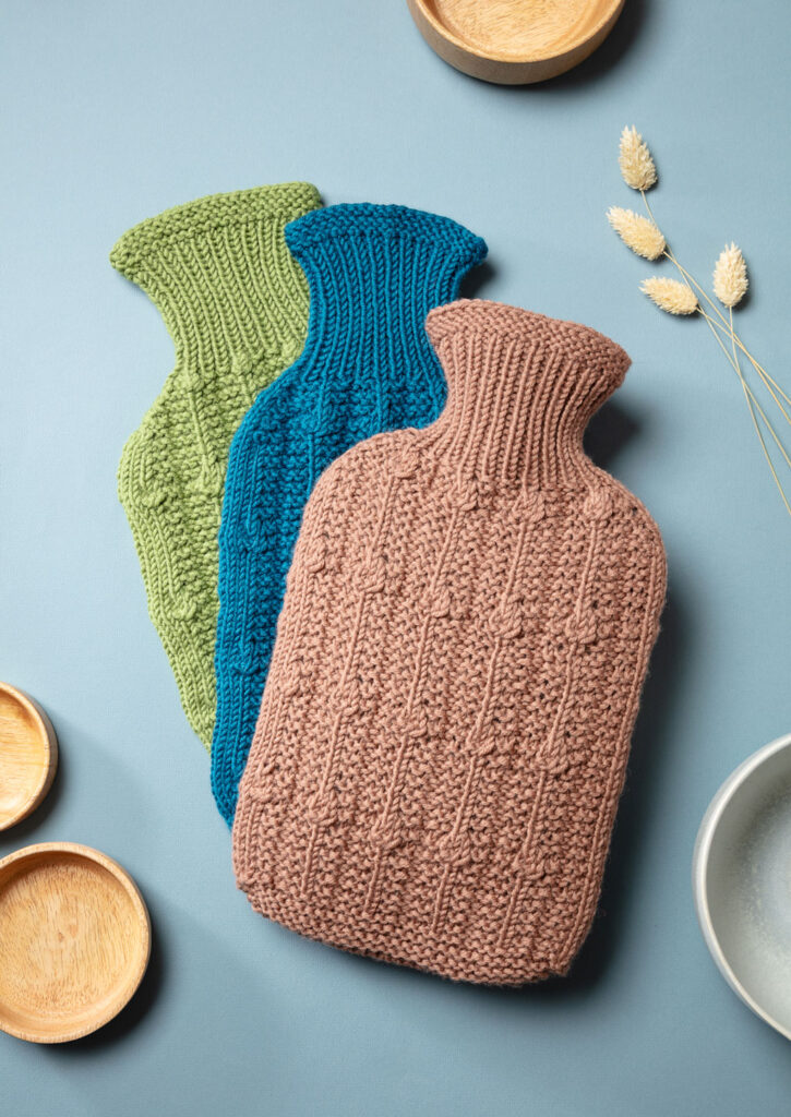 Produktfoto von drei gestrickten Wärmflaschenbezügen mit Muster als Flatlay fotografiert mit natürlichen Deko-Elementen